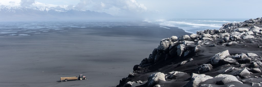 A tractor in the distance set against a rugged black sand coastline and snow-capped mountains